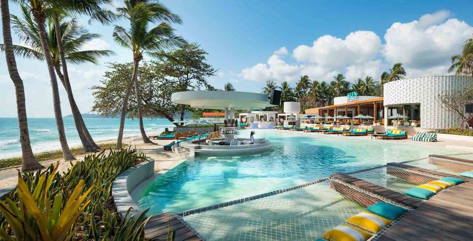 A beachfront pool with a swim-up bar, colourful lounge chairs, and palm trees, overlooking the ocean