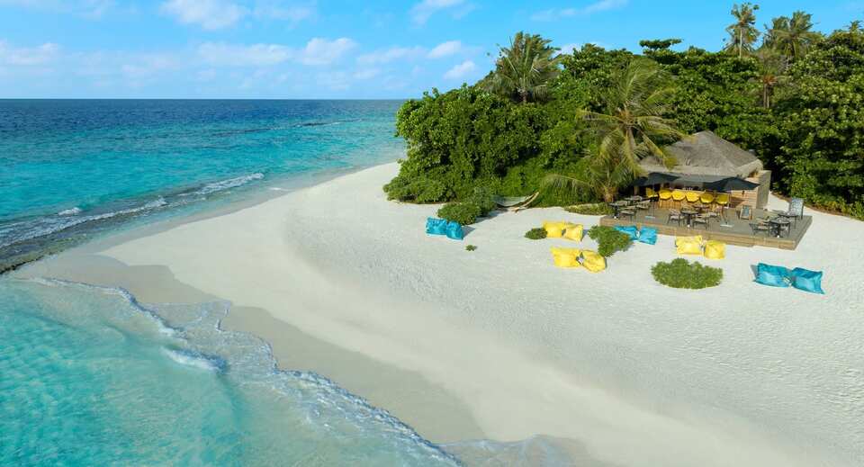 A beachside shack with yellow and blue bean bags on white sand, surrounded by lush greenery, overlooking turquoise Indian ocean